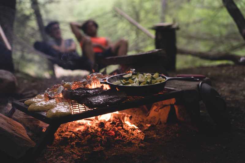 A grate sits over a campfire while some food is cooking, and two individuals in a hammock are seen in the background