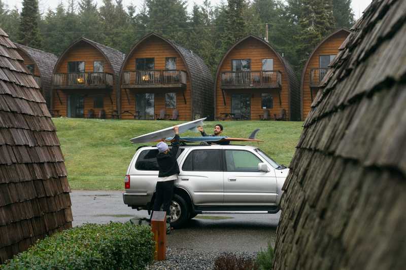 Two people loading a surfboard on top of a SUV, with a row rustic cabins in the background