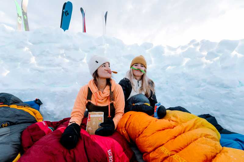 Two females holding up sporks as mustaches in their sleeping bags inside of a snow shelter eating a prepared dehydrated meal