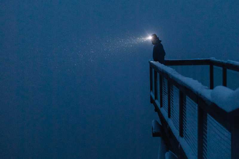 Person on a lookout deck peering into a void of snow flakes illuminated by a headlamp