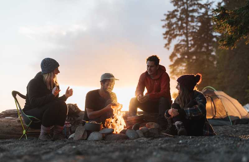 Four friends gathered around a campfire on a beach with a tent in the background